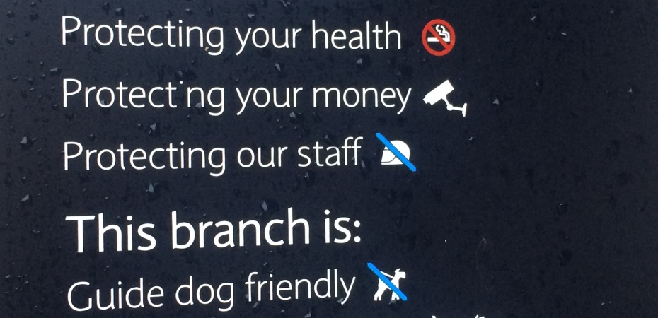 A rain-splashed sign outside a bank reading: "We care about:/Protecting your health/Protecting your money/Protecting our staff/This branch is:/Guide dog friendly/Accessible to all". The sign features symbols of a hearing loop, a wheelchair user, a sight-impaired eye and hands signing.